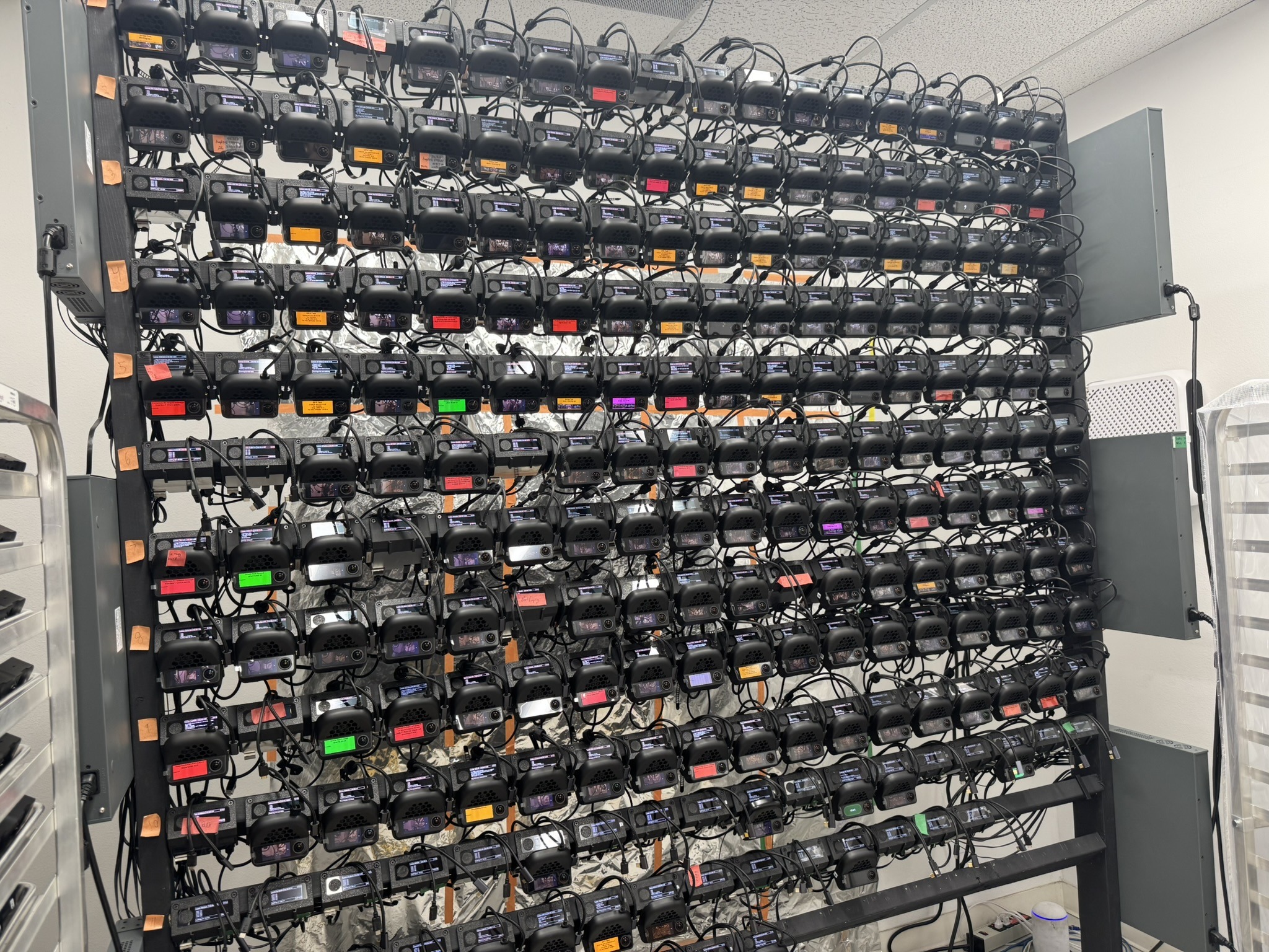 A floor-to-ceiling rack holding hundreds of comma devices, each with its screen lit, labeled with colored tape and wired into rows