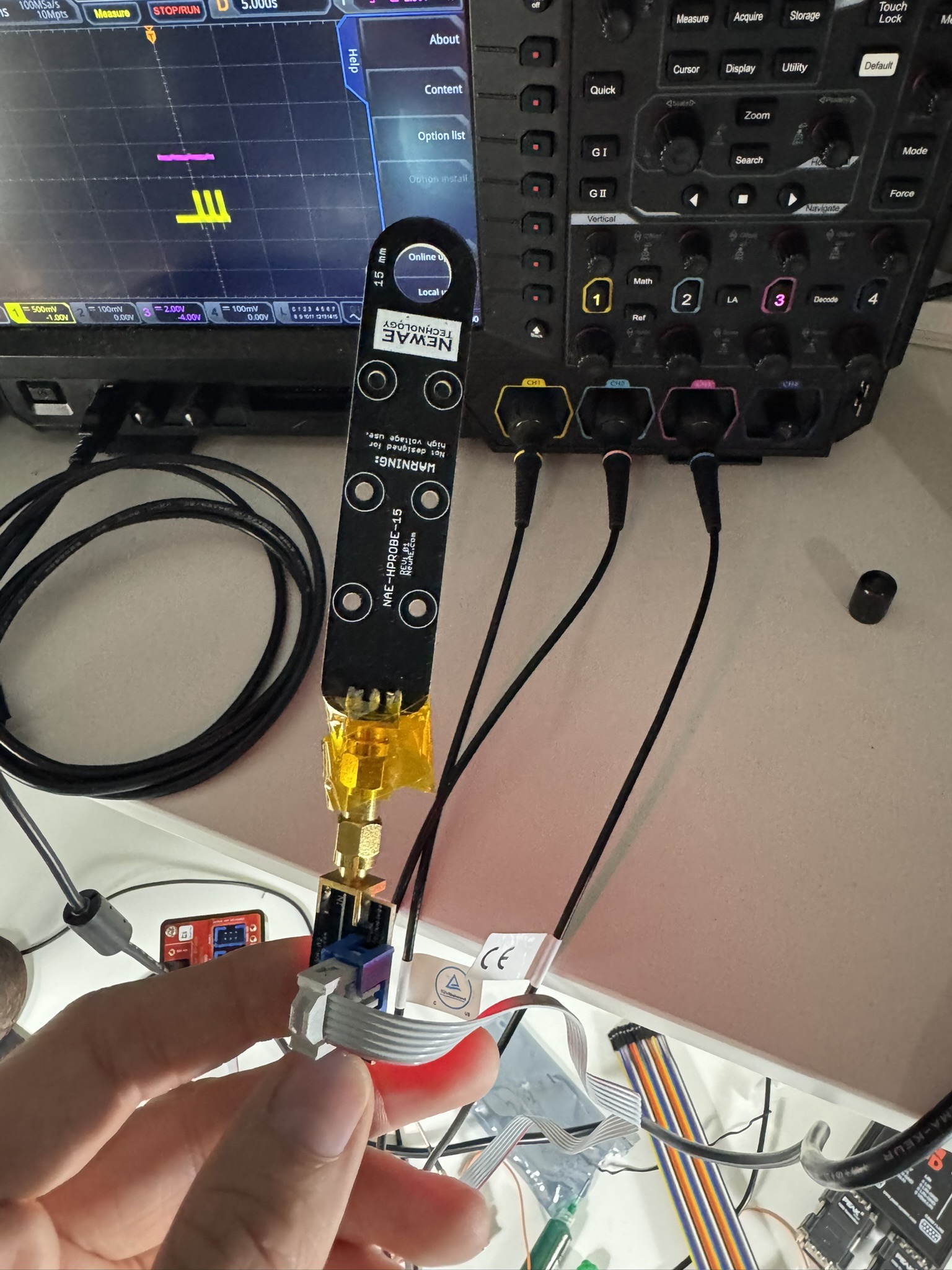 Hand holding the NewAE 15 mm H-field probe, its SMA end wrapped in kapton tape, in front of a Rigol oscilloscope with three channels connected and a CAN frame on screen